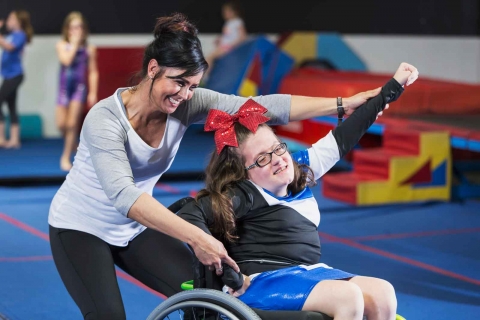 a young teen taking part in wheelchair cheerleading, being assisted by an adult woman 
